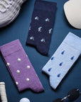 Three pairs of novelty golf socks laid flat on a dark blue surface: one navy pair with small golfer illustrations, one purple pair with white golf balls, and one light blue pair with blue golf balls. A white sneaker, beige cap, golf ball, sunglasses, and grip tape are placed around them as styling props.