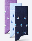 Three pairs of patterned socks displayed in a vertical stack on a light gray background. The front pair is dark navy blue with a repeating golf club and ball player motif, the middle pair is light blue with blue and white golf ball and club motifs, and the back pair is purple with white golf ball and green stem motifs.
