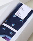 A pair of navy blue socks with a golf-themed print, presented inside a white gift box. The socks feature illustrations of golfers and are branded with a white paper tag that reads 'THE PUTTER by THE LONDON SOCK EXCHANGE'.