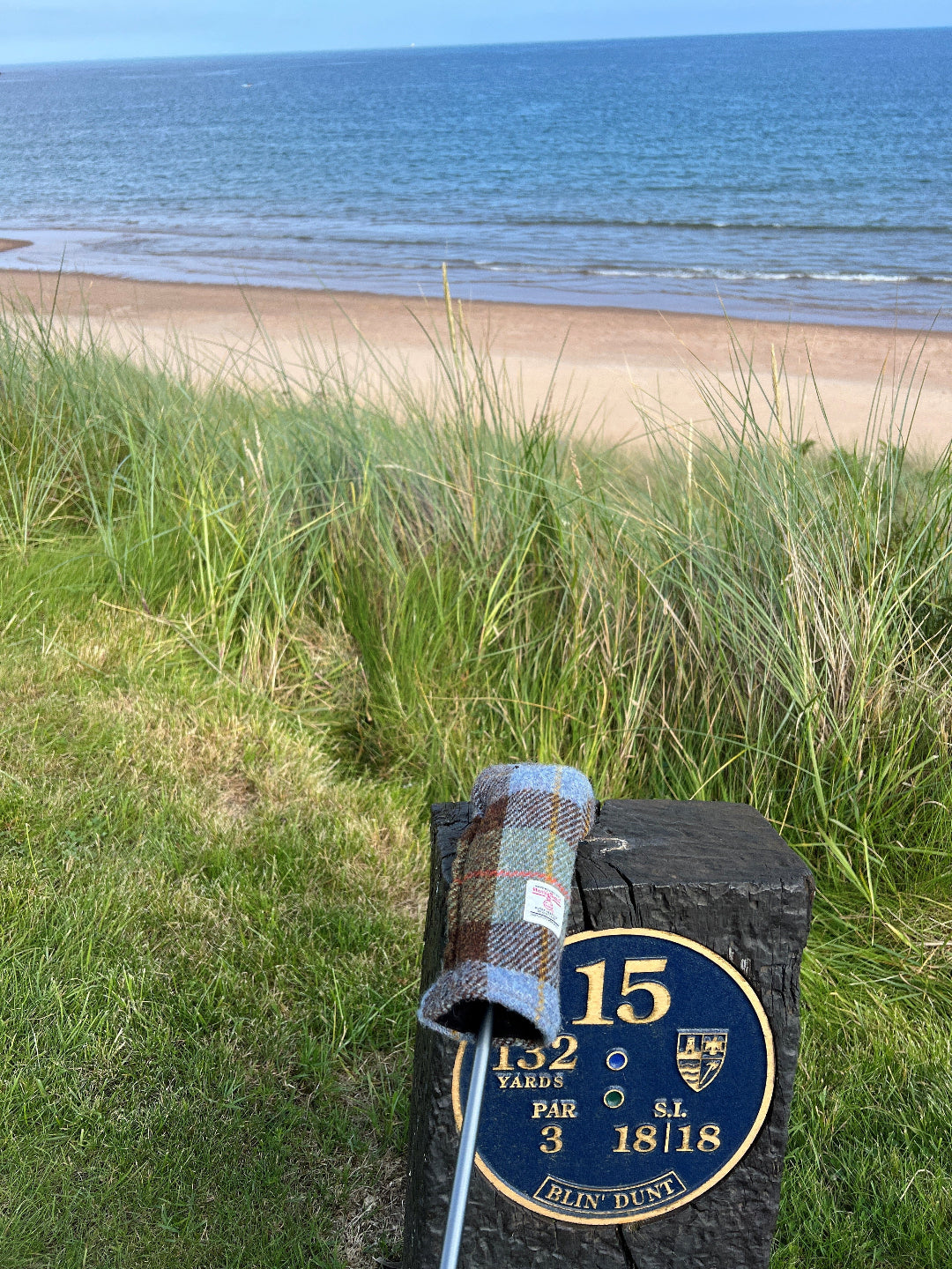 A golf club head with a blue and brown plaid cover rests on a black marker sign at the front of a beach dune, with grass, sand, and the ocean in the background.