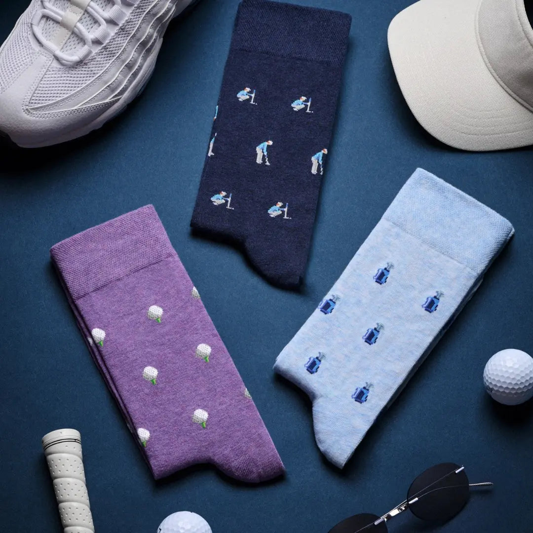 Three pairs of novelty golf socks laid flat on a dark blue surface: one navy pair with small golfer illustrations, one purple pair with white golf balls, and one light blue pair with blue golf balls. A white sneaker, beige cap, golf ball, sunglasses, and grip tape are placed around them as styling props.