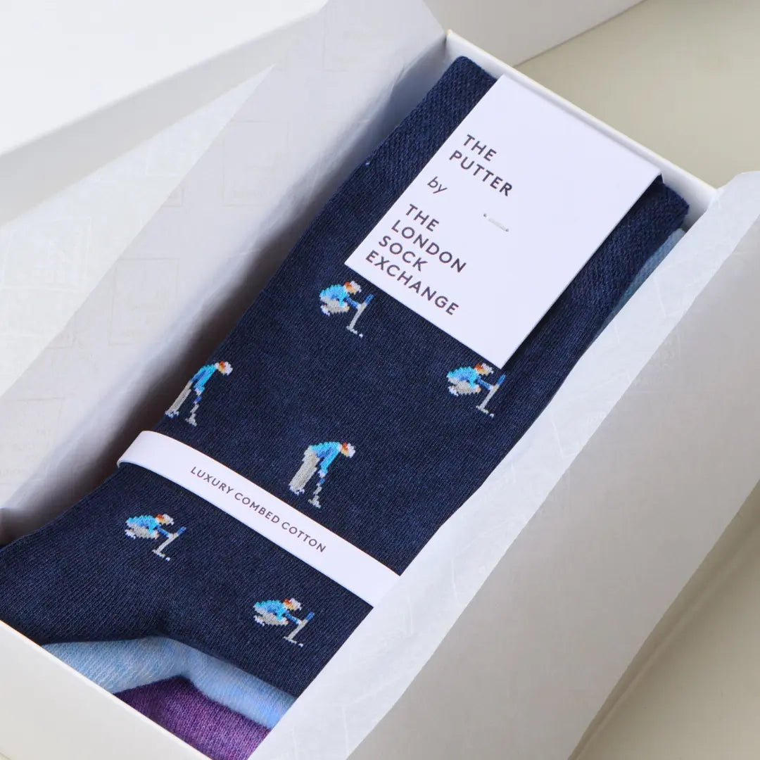 A pair of navy blue socks with a golf-themed print, presented inside a white gift box. The socks feature illustrations of golfers and are branded with a white paper tag that reads 'THE PUTTER by THE LONDON SOCK EXCHANGE'.