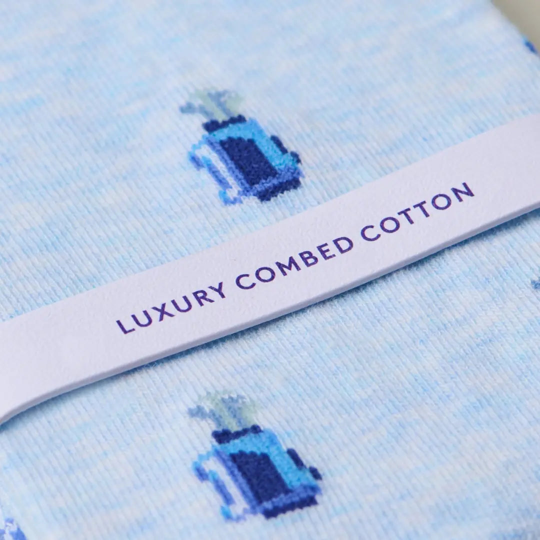 Close-up of light blue knit fabric with a repeating small blue and navy bottle motif and a white paper band across the center printed with the words 'LUXURY COMBED COTTON' in purple.