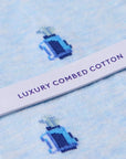 Close-up of light blue knit fabric with a repeating small blue and navy bottle motif and a white paper band across the center printed with the words 'LUXURY COMBED COTTON' in purple.
