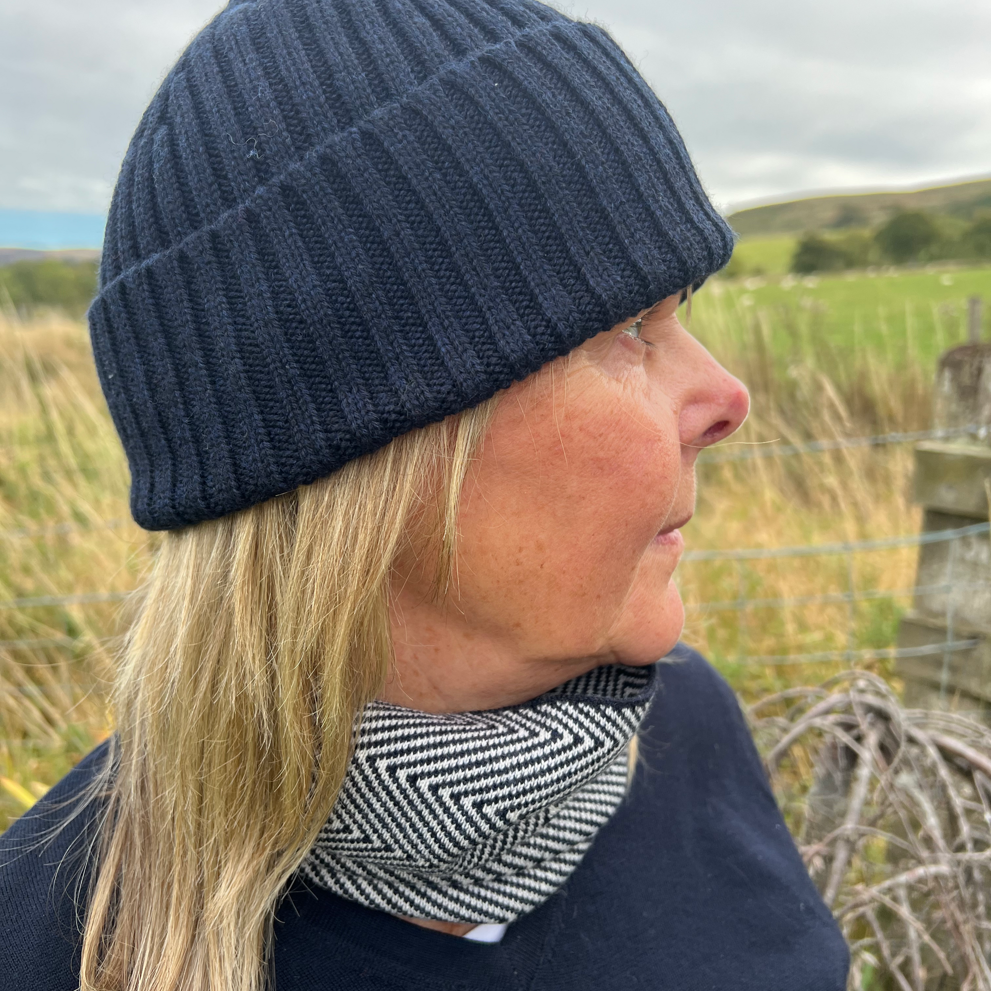 Person wearing a navy knit beanie and patterned scarf outdoors with grass and sky in the background