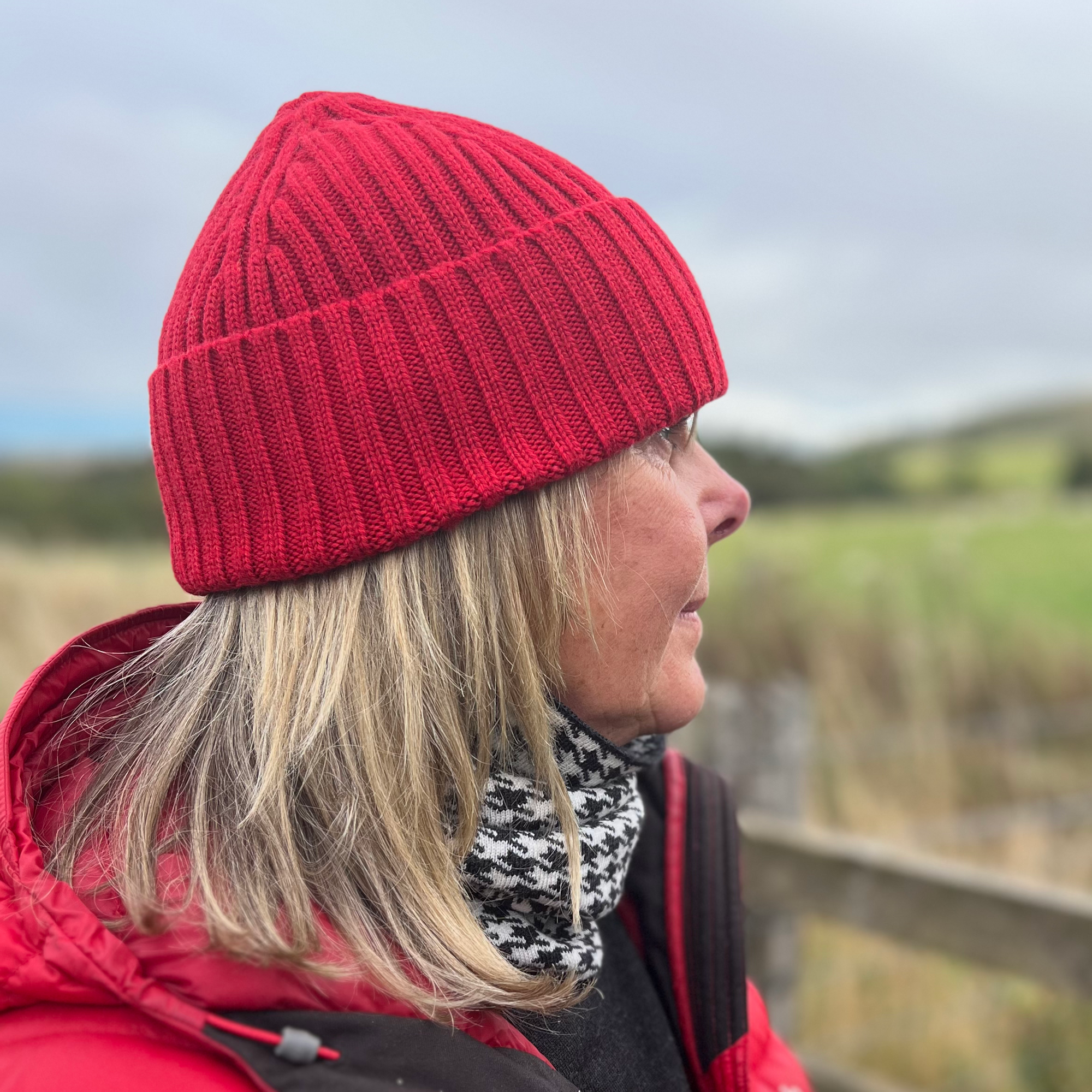 Person wearing a red beanie and jacket standing in a field with a wooden fence and grassy landscape.
